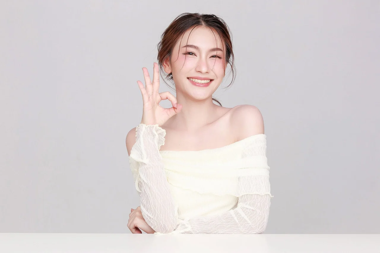 A smiling young woman making an OK gesture with her hand, wearing a white off-shoulder top.