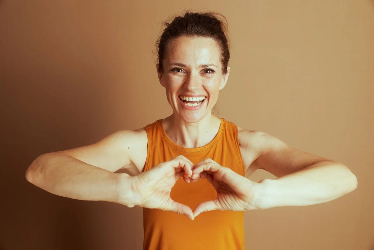 A smiling woman in an orange tank top making a heart shape with her hands.