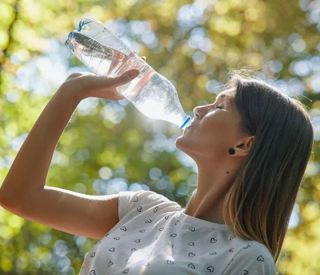 A woman drinking water from a plastic bottle outdoors on a sunny day.