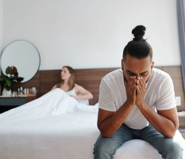 Lying man sitting on bed with woman resting in background in bedroom setting.