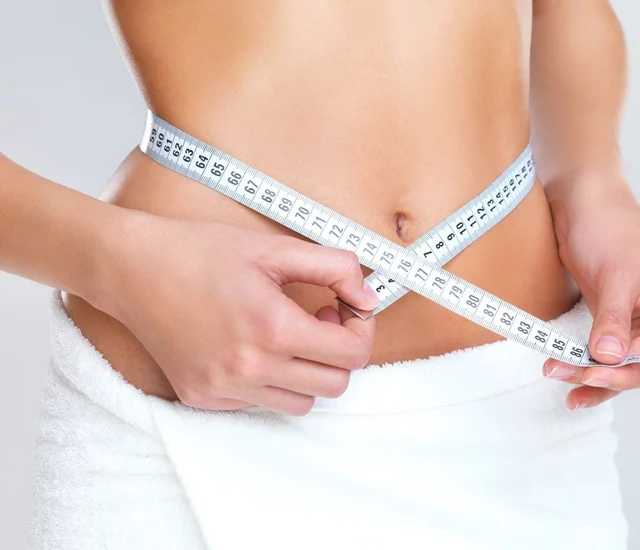 A woman measuring her waist with a tape measure, focusing on abdominal area.