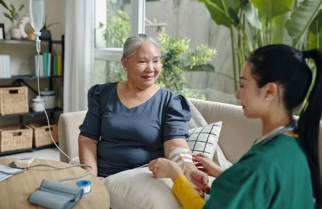 An elderly woman receiving an IV drip while smiling and talking with a healthcare professional.