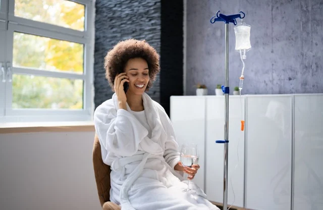 A woman smiling and talking on the phone while sitting in a hospital gown next to an IV drip.