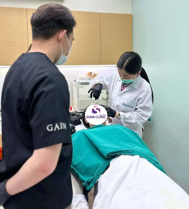 A woman in medical attire performs a procedure on a patient lying in a clinic bed, with a man observ.
