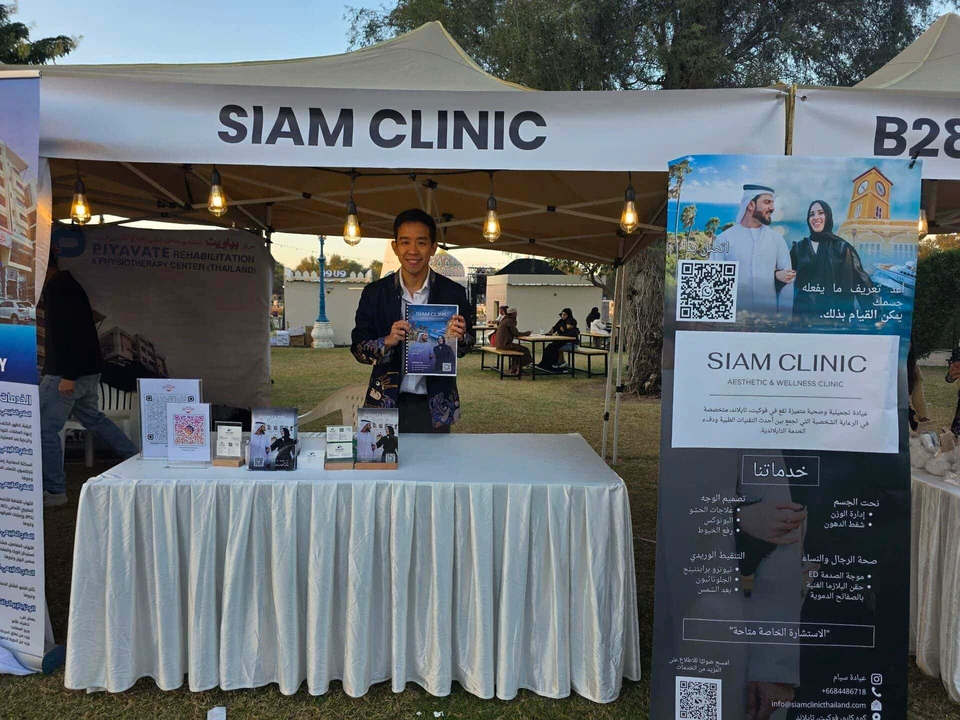 A smiling man standing behind a table at an outdoor booth with a "SIAM CLINIC" banner, promotional m.