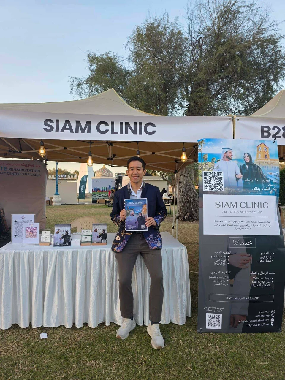A smiling man standing in front of a Siam Clinic booth at an outdoor event, holding a brochure.