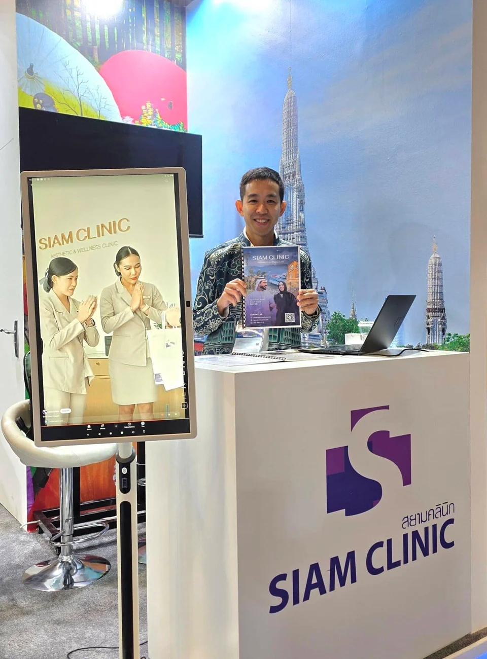 A smiling man standing behind a SIAM CLINIC booth, holding a magazine, with a large digital display.