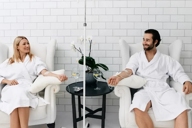 A woman and a man sitting comfortably in white robes, receiving IV therapy in a wellness clinic.