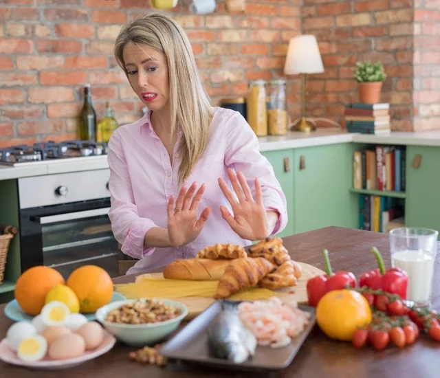 A woman in a pink shirt gesturing with her hands in a kitchen with various foods on the table.