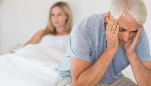 Feeling distressed and upset, man sitting on bed with woman in background.