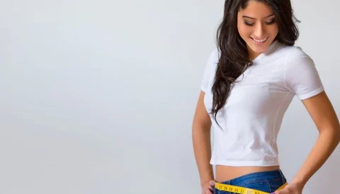 Slim woman measuring her waist with a tape measure, smiling, wearing a white t-shirt and jeans.