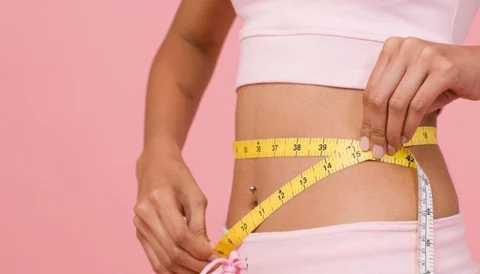 A woman measuring her waist with a yellow tape measure against a pink background.