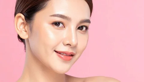 A woman with clear skin and a gentle smile, posing against a pink background.