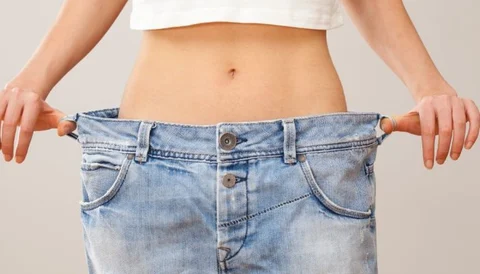 A woman holding out oversized jeans to show a slim waist and flat stomach.