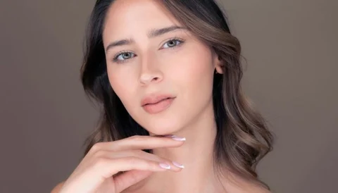 A young woman with clear skin and wavy brown hair posing against a neutral background.