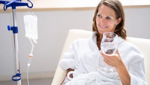 A woman in a hospital gown sitting in a chair, holding a glass of water, with IV drip beside her.