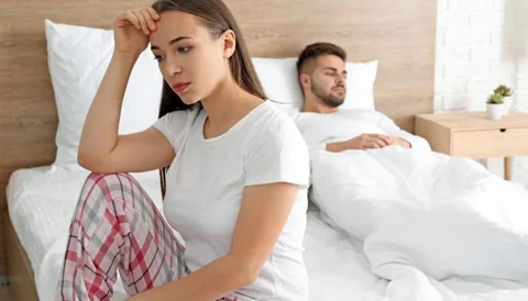 Feeling stressed and tired, woman sitting on bed with man sleeping in background.