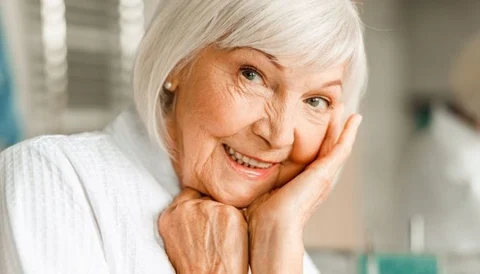 A smiling elderly woman with white hair touching her face, indoors.