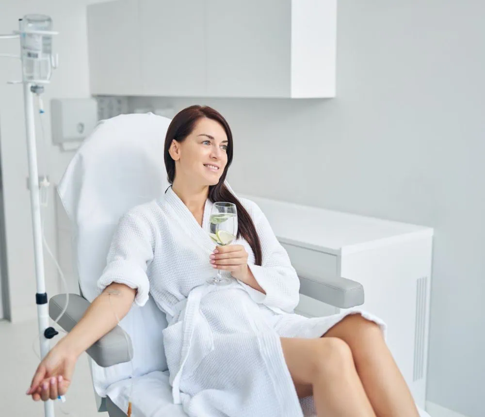 An older woman in a hospital gown sitting in a medical chair, holding a glass of IV fluids.