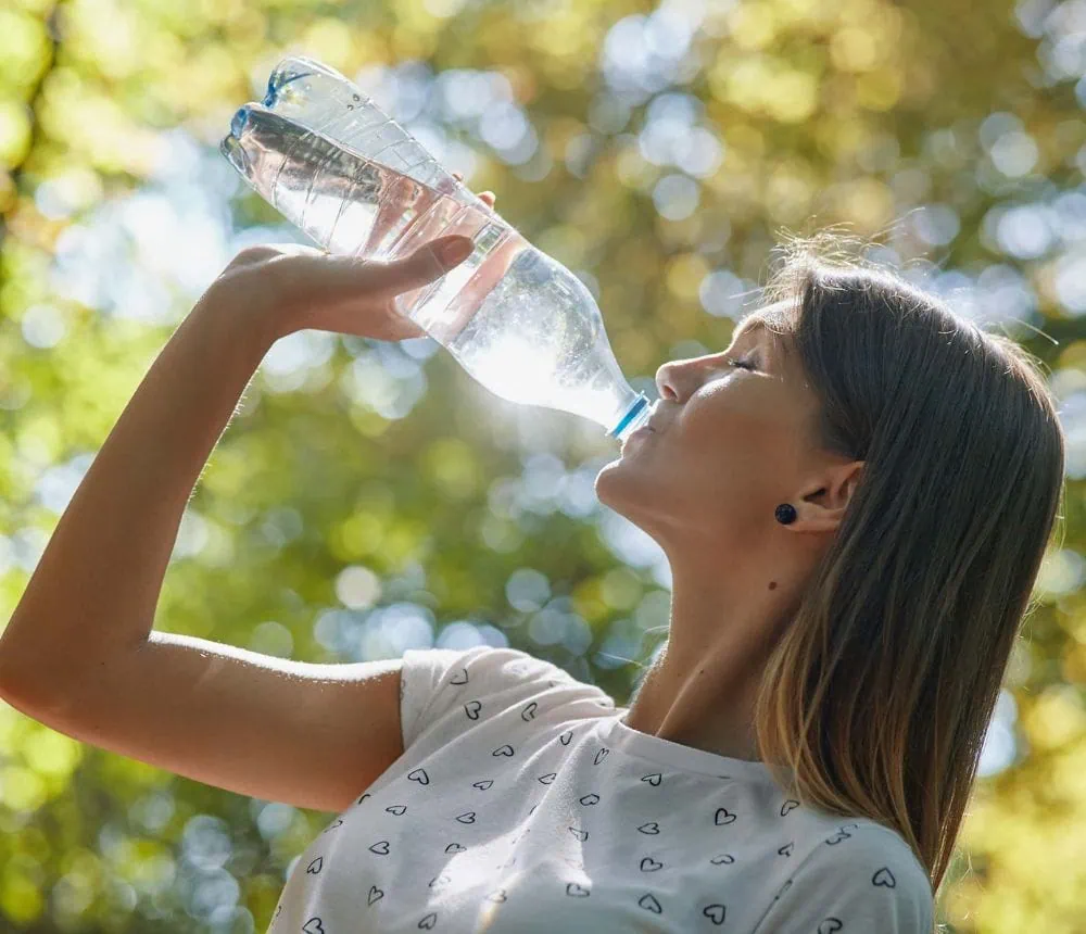A woman drinking water from a plastic bottle outdoors on a sunny day.