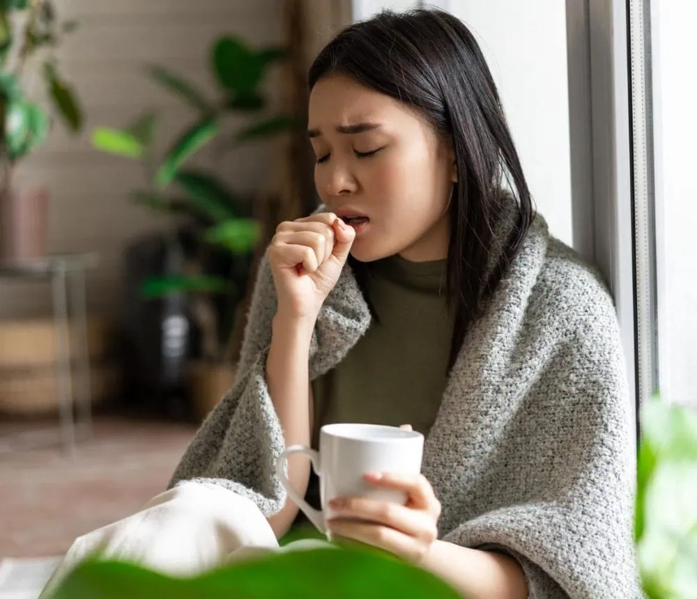 A woman sitting by a window, holding a mug, appears to be coughing or feeling unwell.