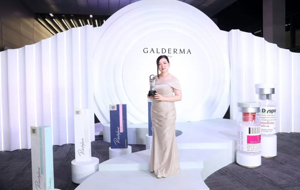 A woman in a beige dress holding an award on a stage with a "Galderma" backdrop and product displays.