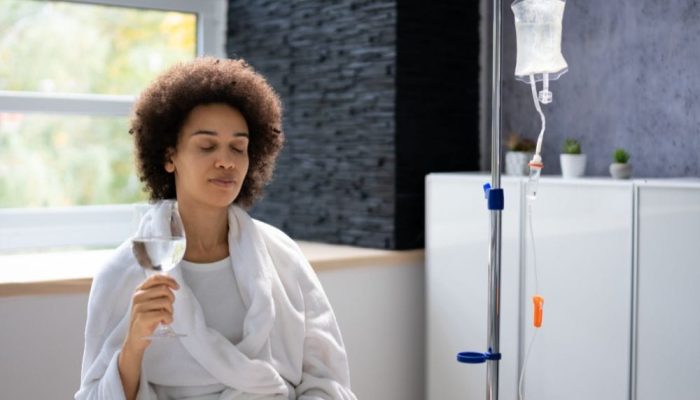 A woman with curly hair sitting in a hospital room, holding a glass of water near an IV stand.