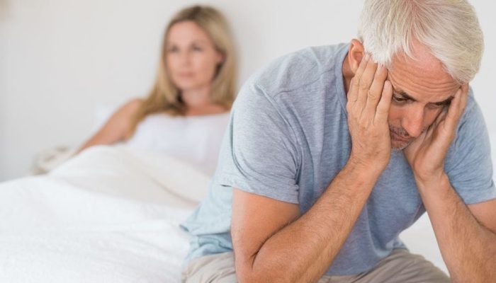 Feeling distressed and upset, man sitting on bed with woman in background.