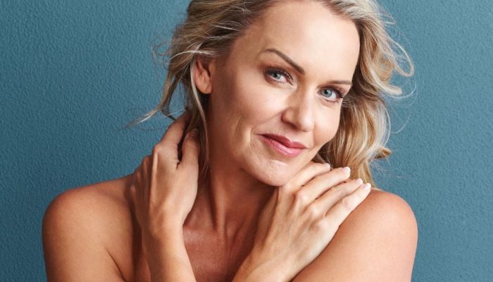 A smiling woman with blonde hair touching her neck against a teal background.