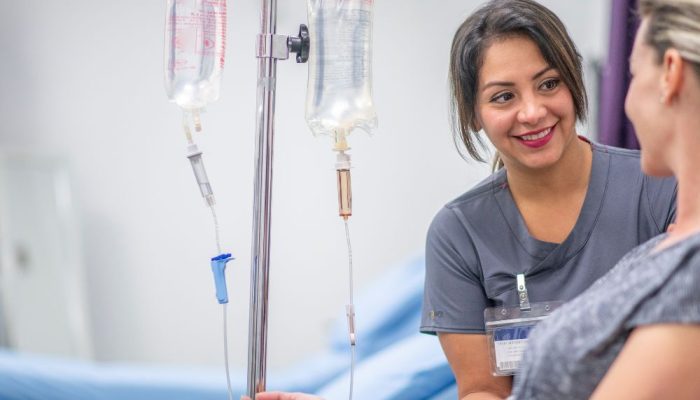 An IV drip connected to a patient in a hospital room with a nurse smiling and talking to her.
