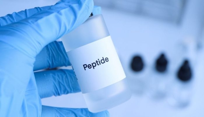 A detailed image of a hand in a blue glove holding a small white bottle labeled "Peptide" with a dro.