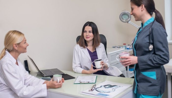 A woman in a gray medical uniform and stethoscope talks to two women in white coats, one holding a m.