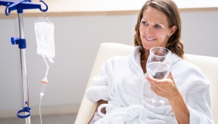A woman in a hospital gown sitting in a chair, holding a glass of water, with IV drip beside her.