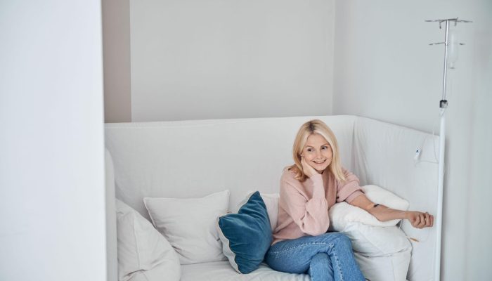 A smiling woman sitting on a white medical couch with an IV drip stand nearby.