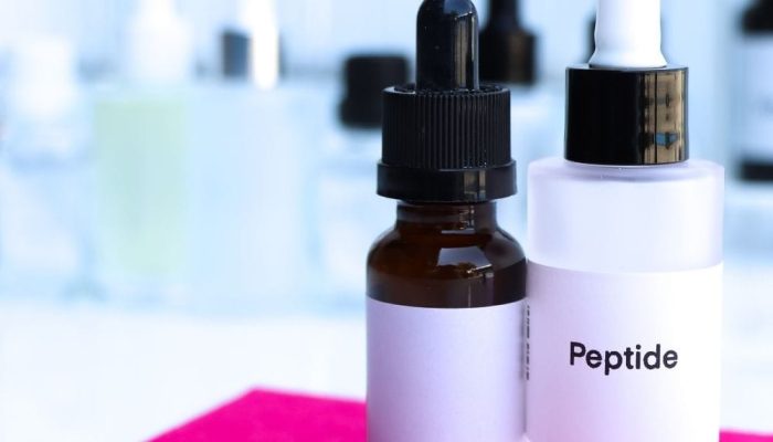 A dropper bottle labeled "Peptide" on a pink cloth with blurred bottles in the background.
