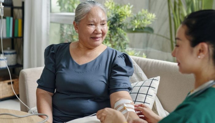 A woman receiving an IV infusion from a healthcare professional in a bright, cozy room.