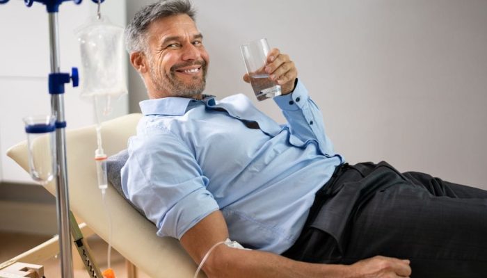A man lying on a medical chair receiving an IV infusion, smiling and holding a glass of water.